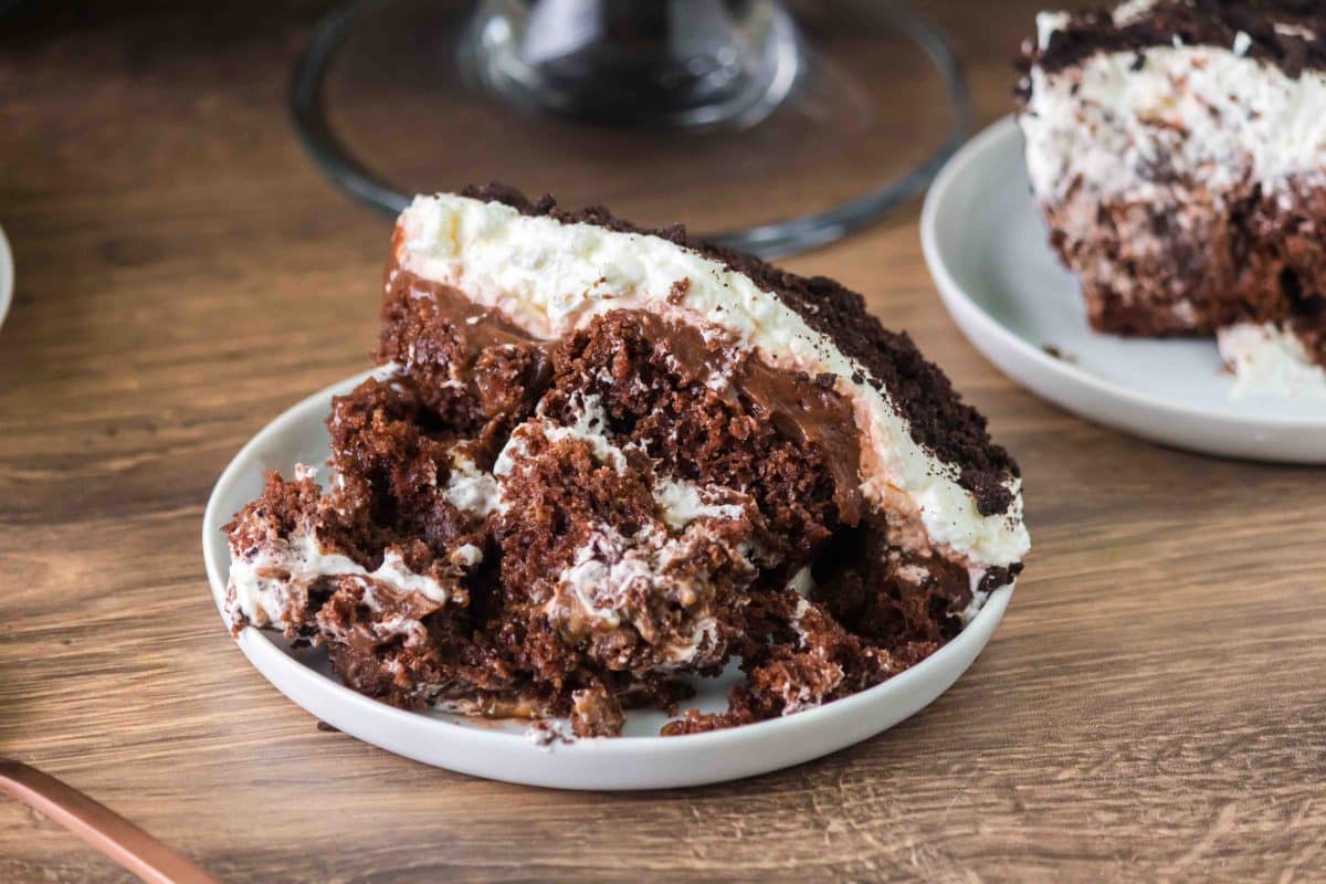 A large serving of a chocolate pudding trifle dessert on a white plate showing layers of chocolate cake, pudding, whipped cream and oreo cookie crumbs. Hostess At Heart