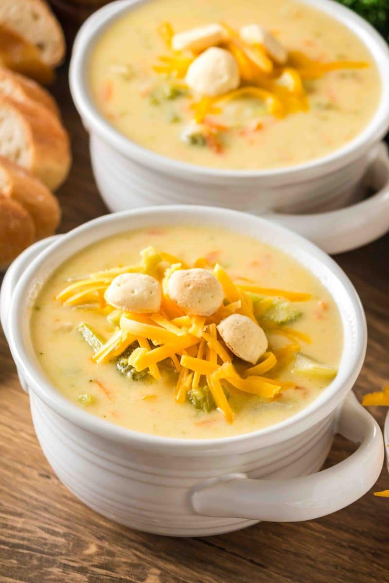 Angled shot of creamy broccoli cheese soup in a white bowl topped with shredded cheddar and crackers. Hostess At Heart