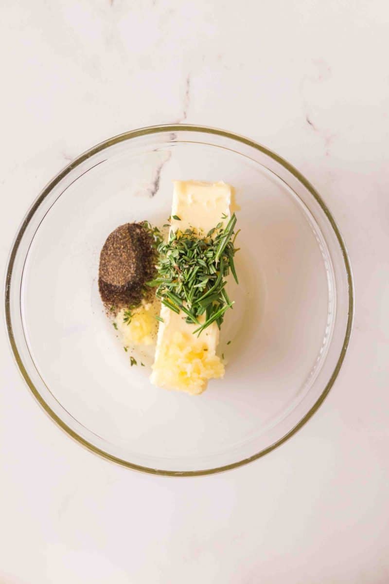 Herb butter ingredients, butter, rosemary, thyme, salt and pepper, in a glass bowl.