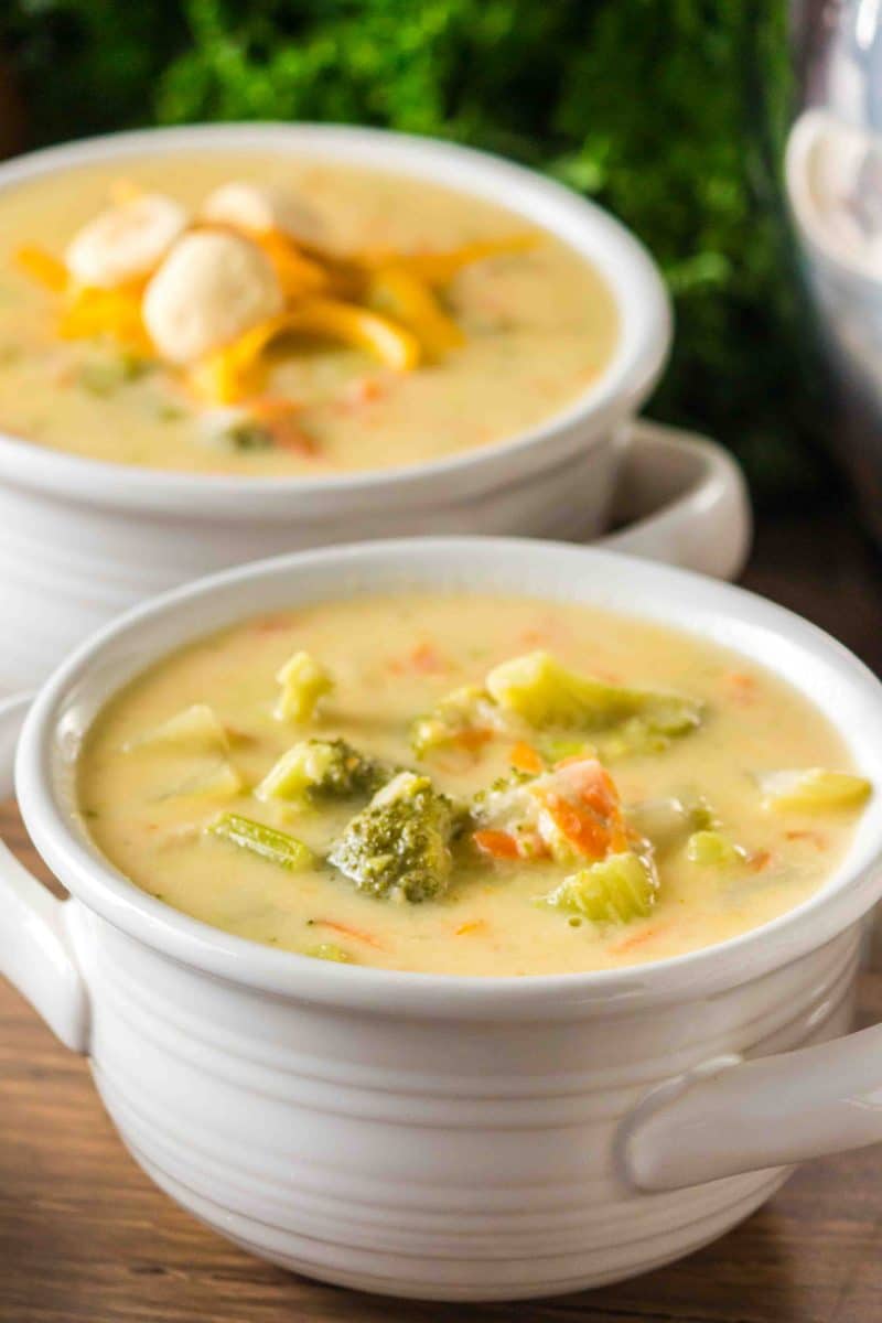 Two bowls of homemade broccoli cheddar soup in a white bowls showing chunks of broccoli, celery, and carrots in a creamy broth. Hostess At Heart