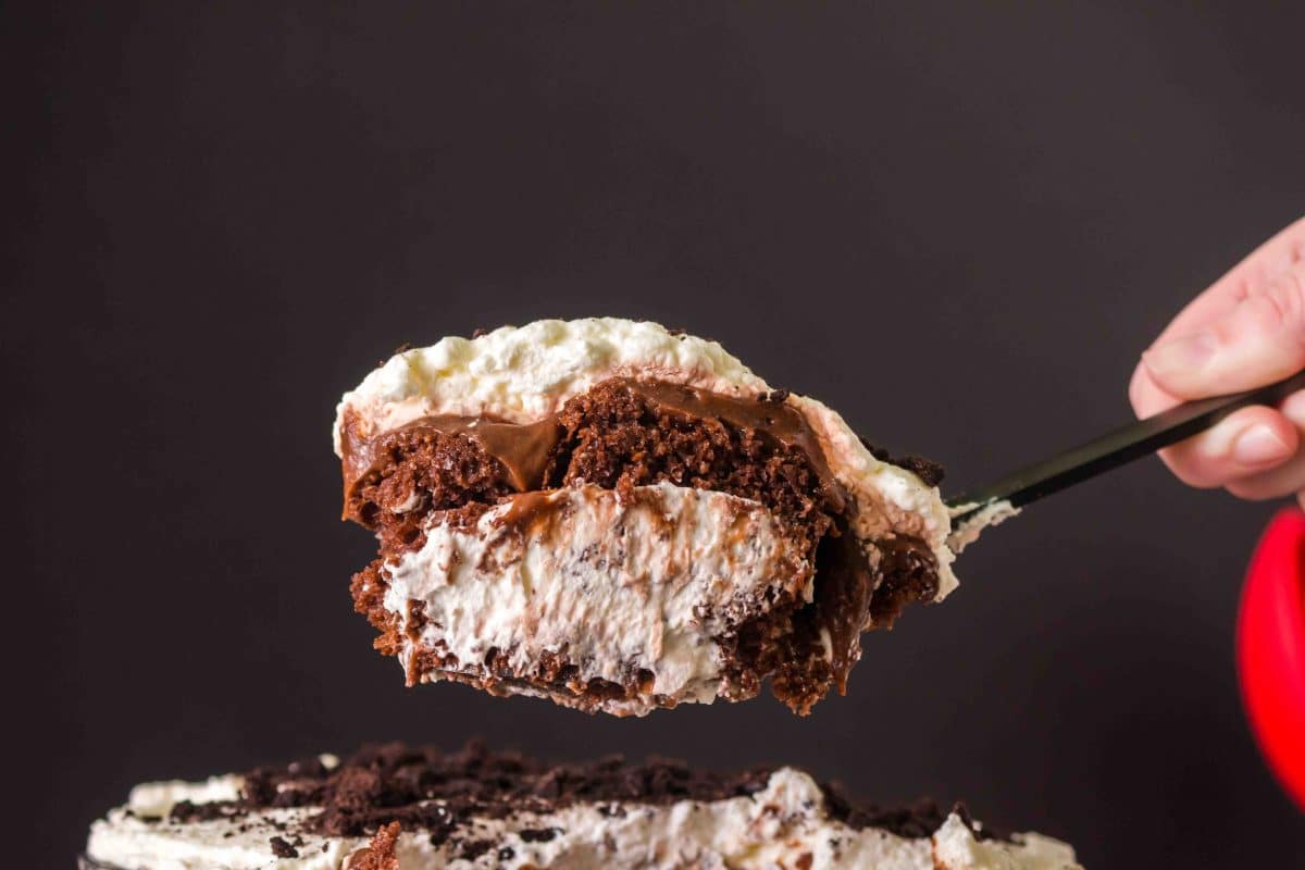 A large spoon lifting a serving of chocolate pudding trifle desert showing layers of chocolate cake, creamy chocolate pudding, sweet whipped cream, and a sprinkle of Oreos. Hostess At Heart