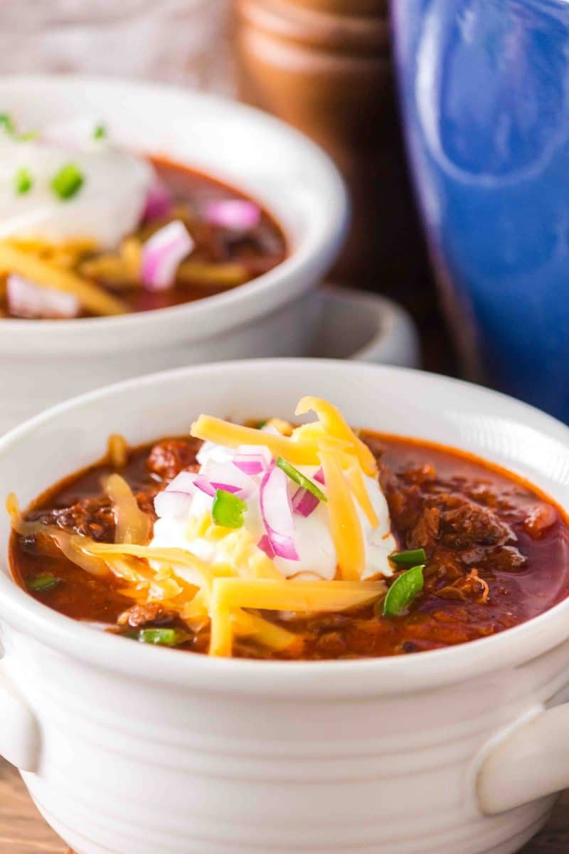 Angled view of white soup bowl filled with brisket chili recipe garnished with sour cream, shredded cheese, red onion, and diced jalapeno. Hostess At heart
