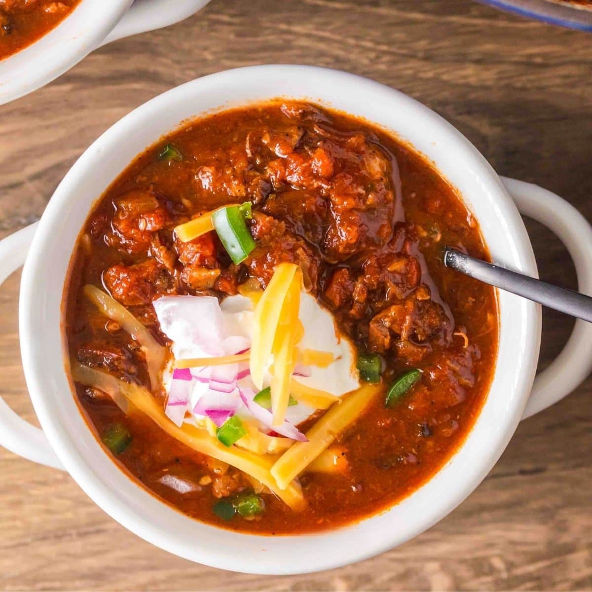Overhead view of a white soup bowl filled with brisket chili garnished with sour cream, shredded cheese, and red onions. Hostess At Heart