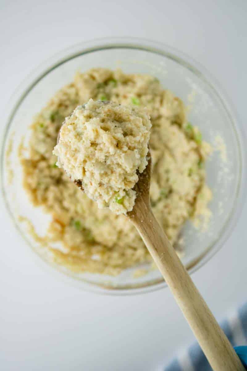 A wooden spoon lifting unbaked cornbread dressing from a mixing bowl showing how thick and moist it it. Hostess At Heart