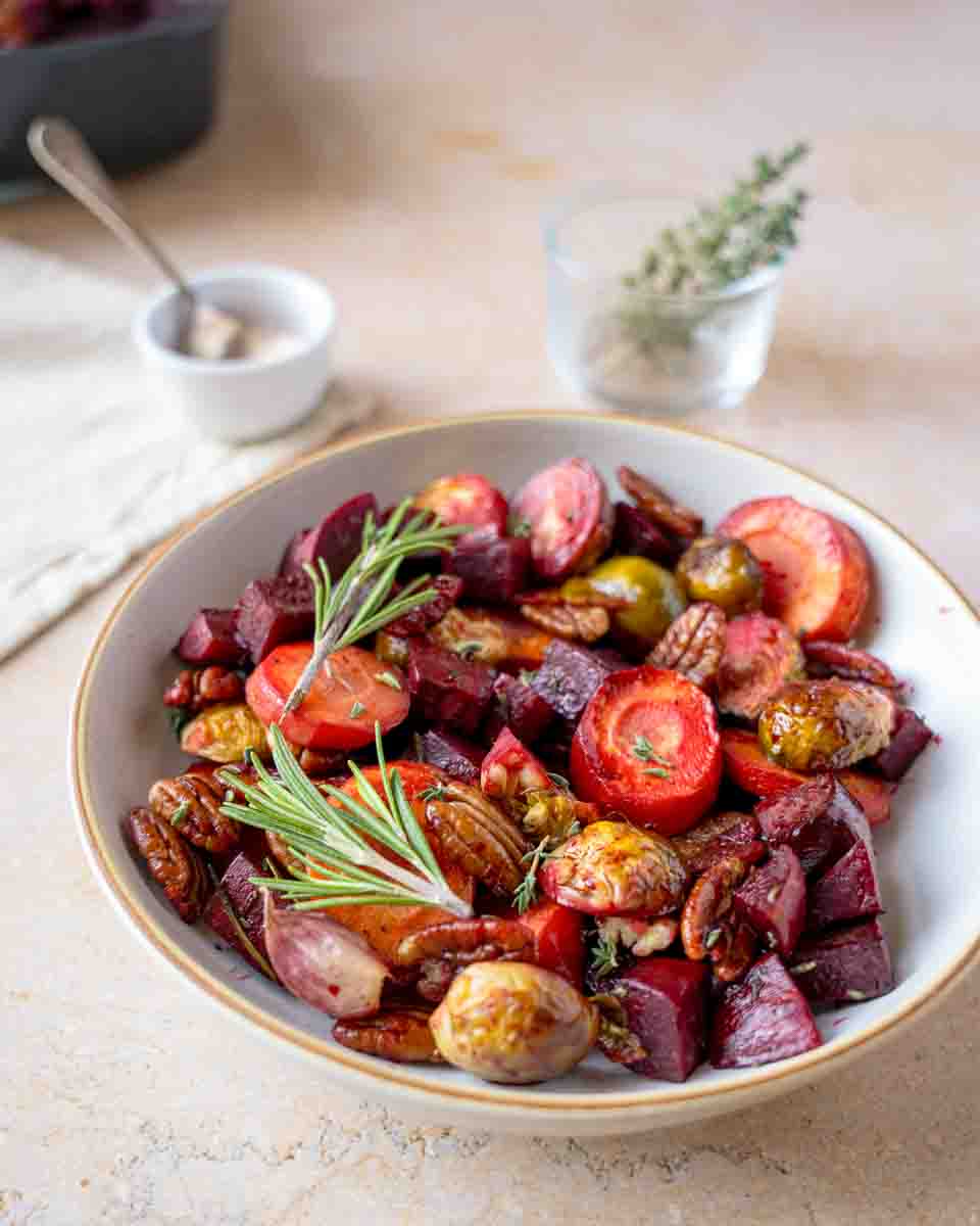 White serving dish filled with roasted beets, Brussels sprouts, and carrots topped with herbs and pecans. Hostess At Heart