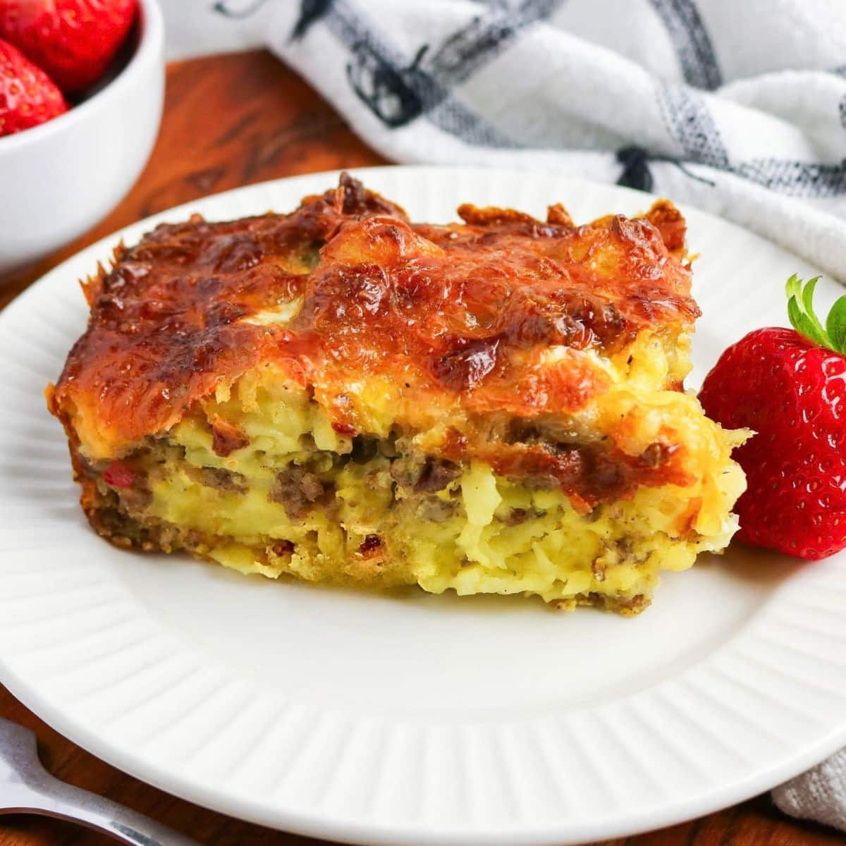 Sideview of a Slice of Breakfast Tater Tot Casserole with sausage, eggs, potatoes, and melted cheese on a white plate. Hostesss At Heart