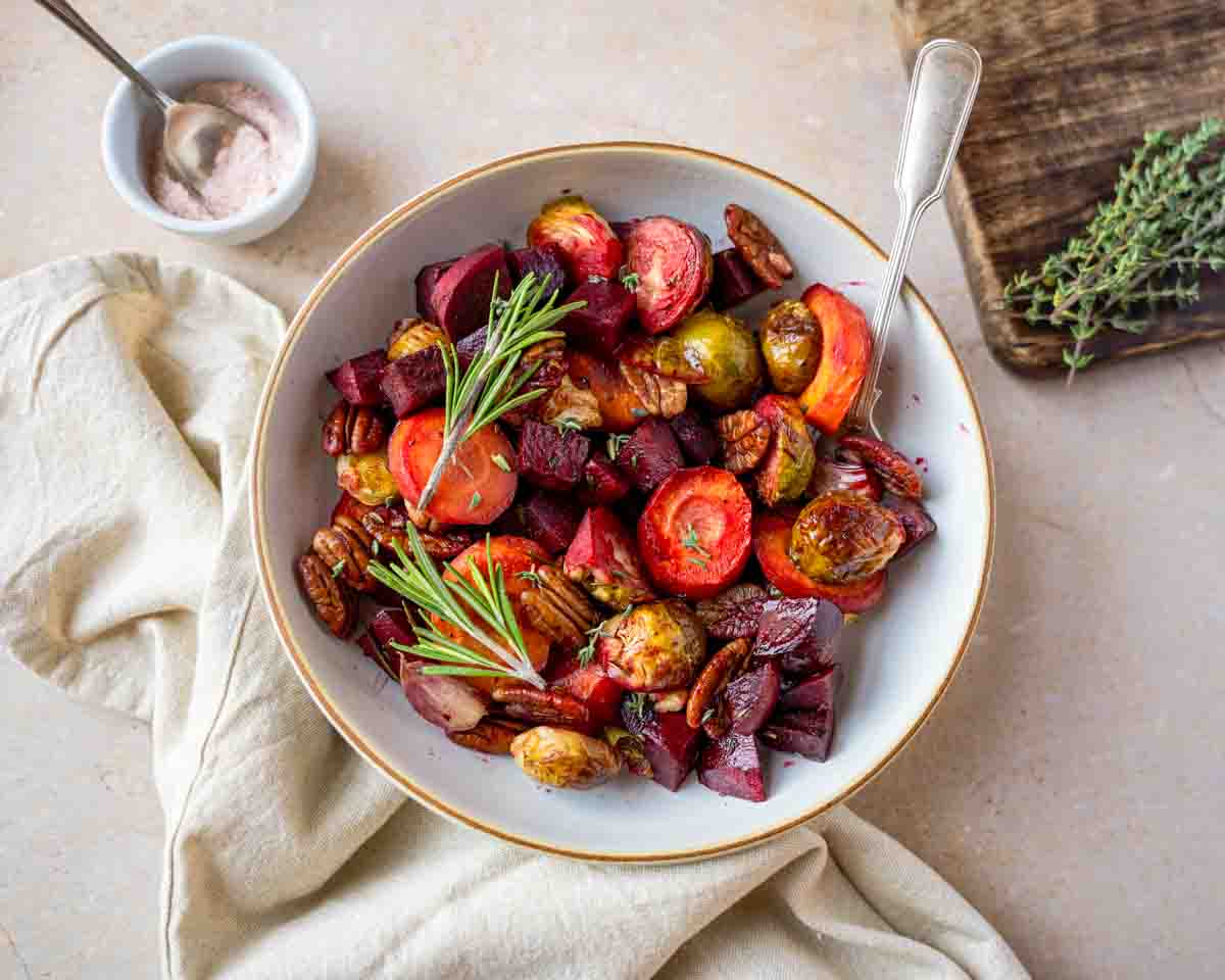 Top down view of a serving dish filled with beautifully roasted holiday vegetables including beets, carrots, Brussels sprouts, and garlic, topped with fresh herbs and pecans. Hostess At Heart