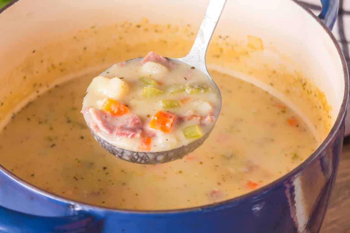 A ladle filled with creamy ham and potato soup with chunky vegetables being lifted from a Dutch oven. Hostess At Heart