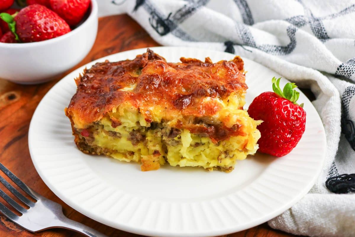 Sideview of a white plate topped with a slice of breakfast tater tot casserole sowing a tender filling of potatoes and sausage under a golden brown crispy cheese crust. Hostess At Heart