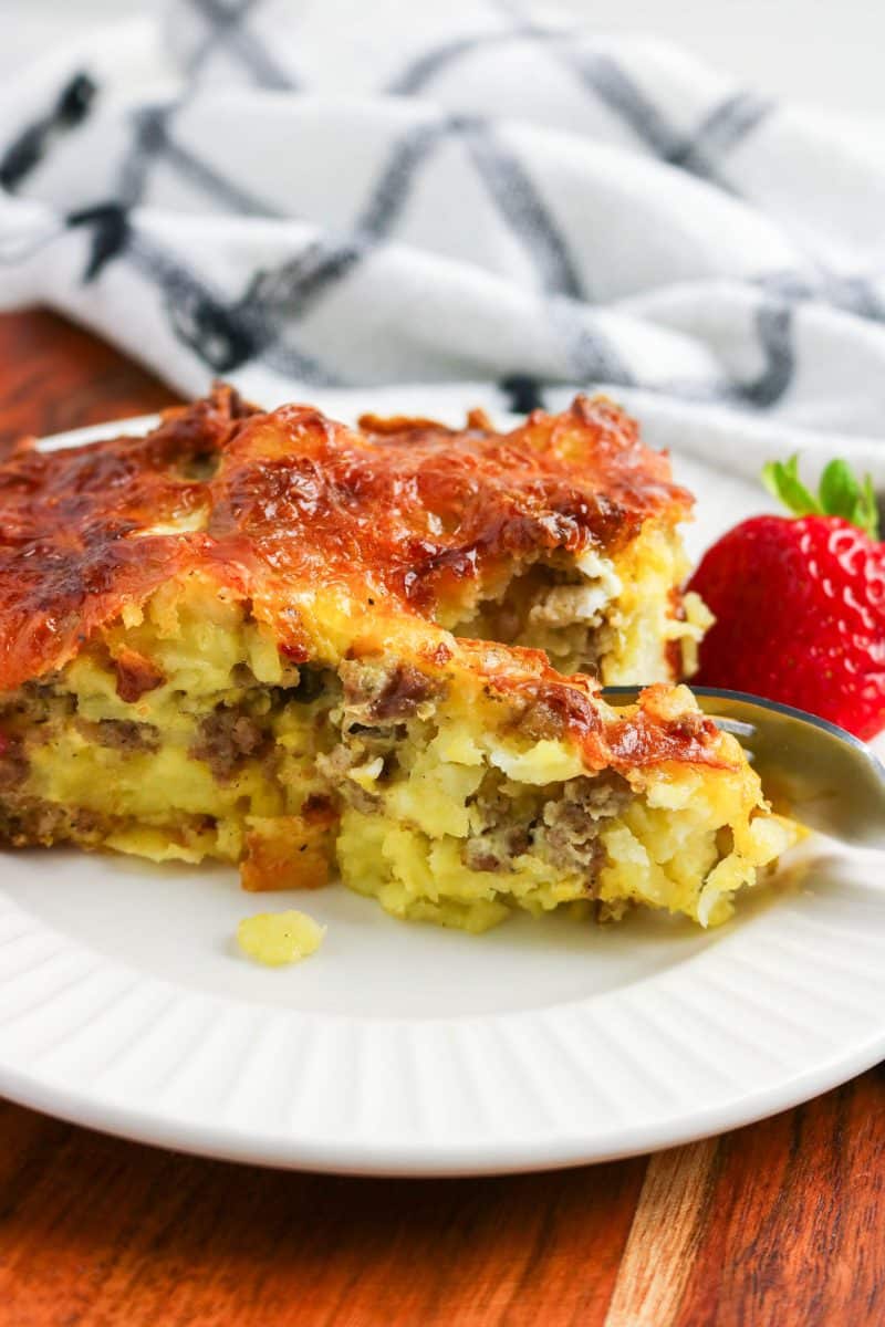 Sideview of a slice of Tater Tot Breakfast Casserole that's being cut with a fork showing a tender interior of sausage and tator tots under a golden crispy baked cheese crust. Hostess At Heart