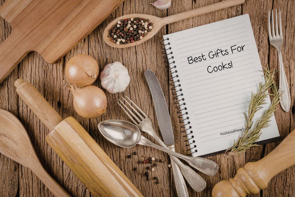 Wooden kitchen tools and a recipe book laying on a wooden cutting board as inspiration for buying gifts for people that love to cook. Hostess At Heart