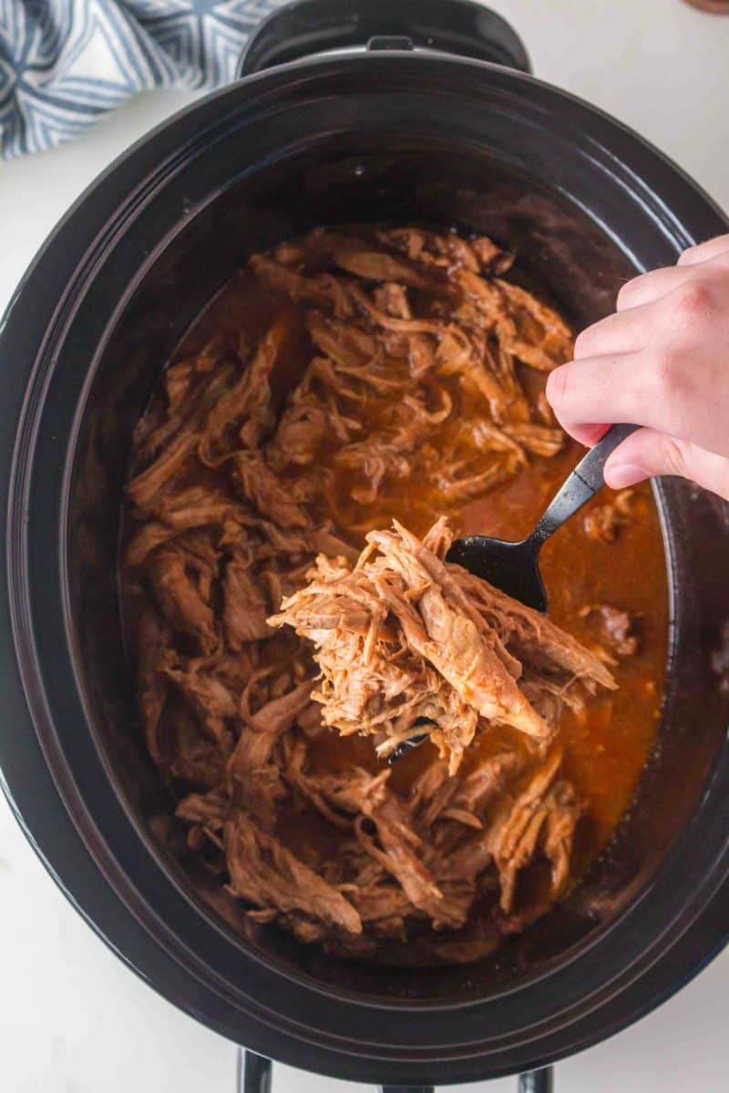 Sauce soaked shredded pork tenderloin being lifted out of a slow cooker. Hostess At Heart